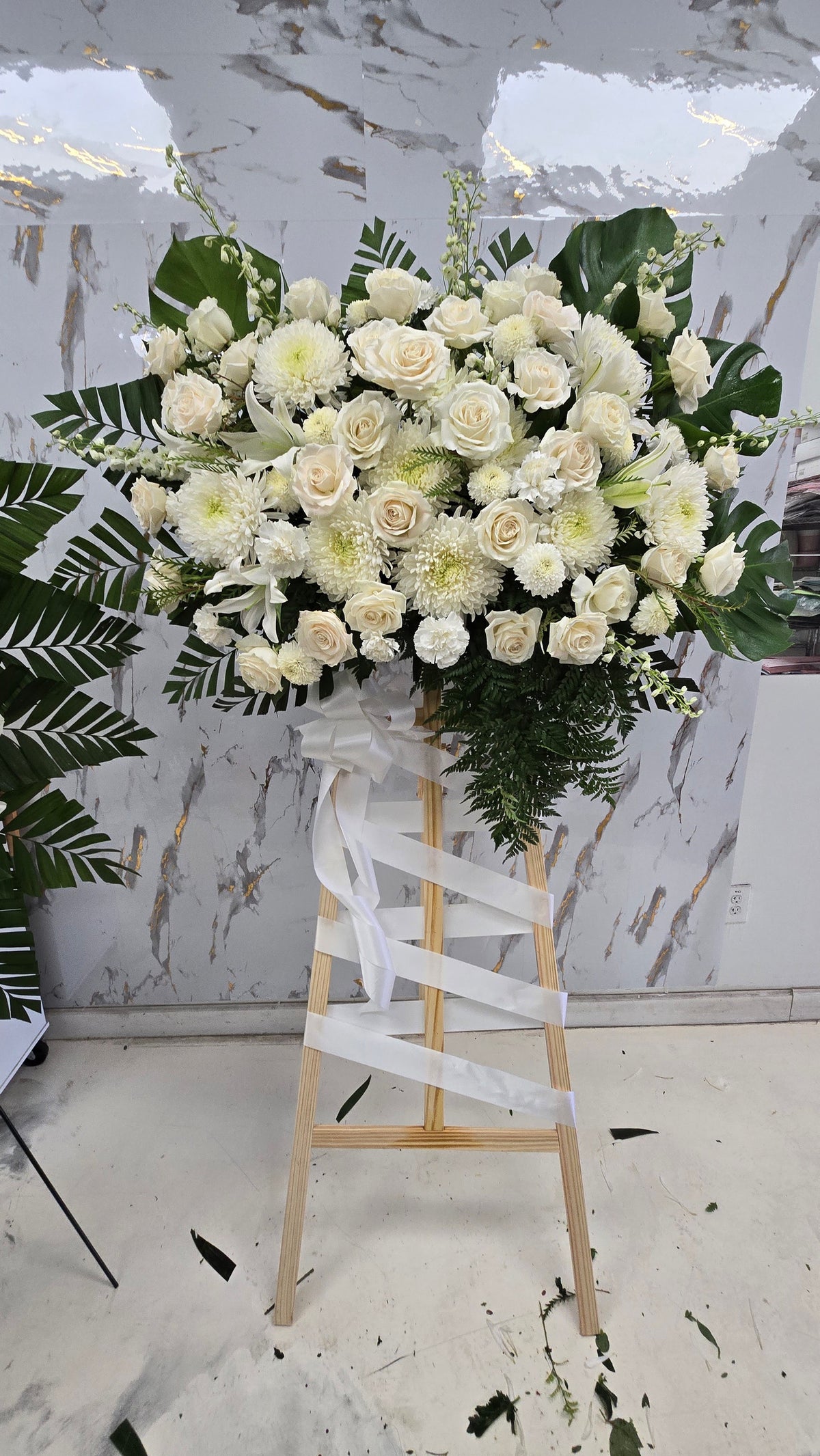Oval white and ivory floral sympathy arrangement with dark green foliage shown upright against a pale wall
