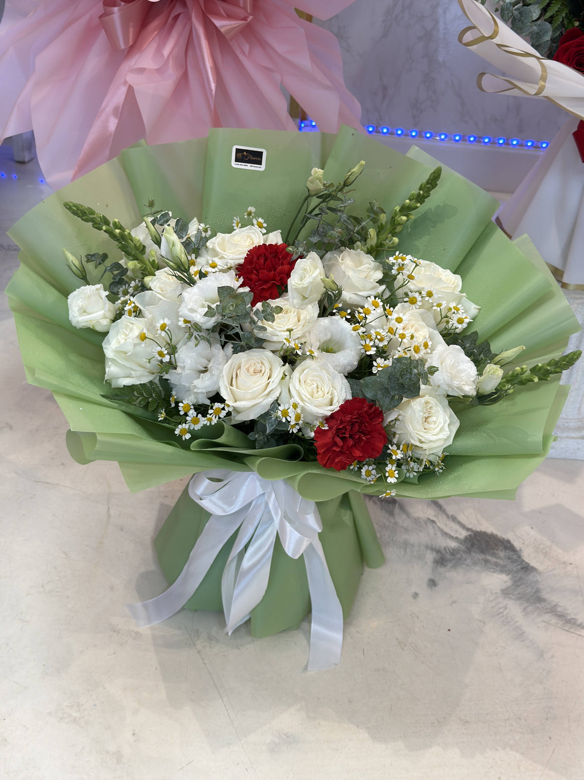 Alternate real view of the white rose bouquet with red accents, small white blooms, sage-green wrapping and a white bow