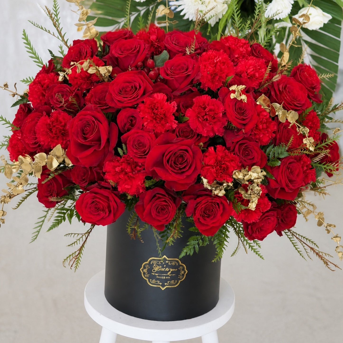 Large red roses and carnations with gold foliage and fern greenery arranged in a black round hatbox