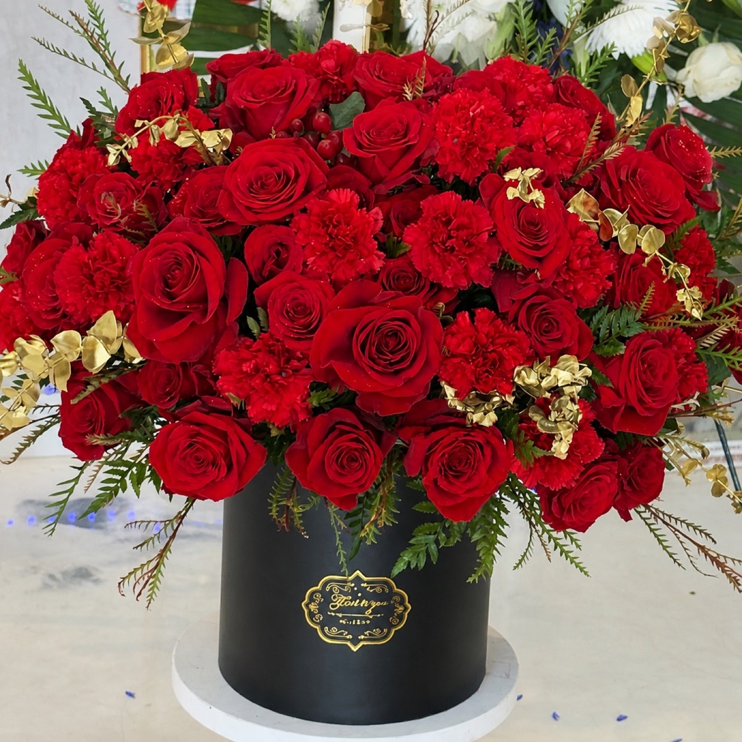 Front view of a large red rose and carnation arrangement with gold foliage in a black round hatbox